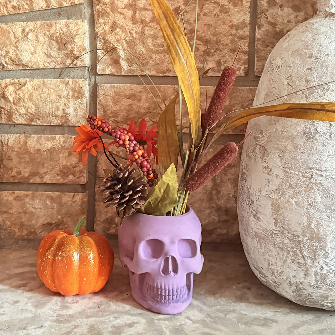 skull shaped candle holder with artificial foliage beside a small decorative pumpkin and large vase on a textured surface