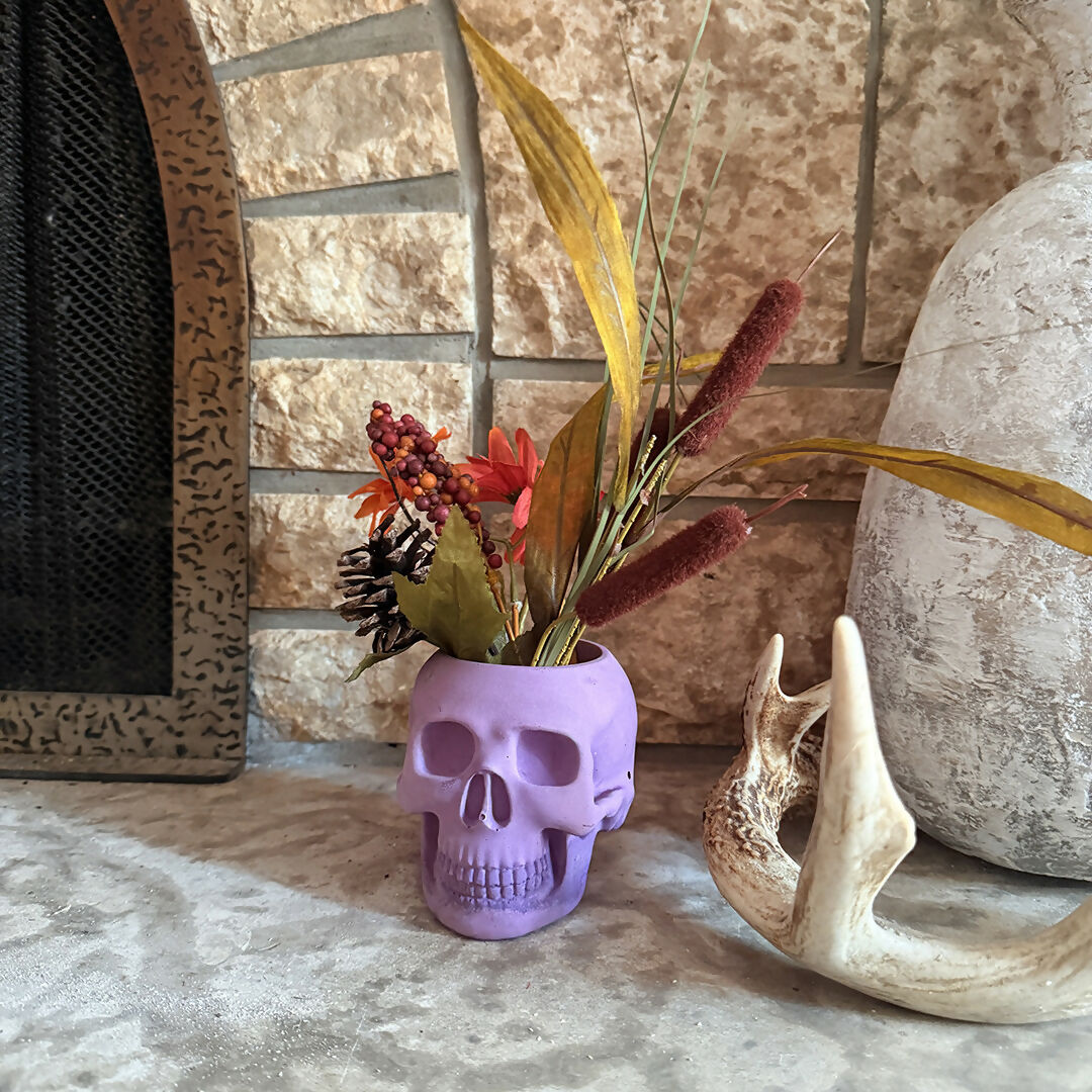skull shaped candle holder with artificial foliage beside a small decorative pumpkin and large vase on a textured surface
