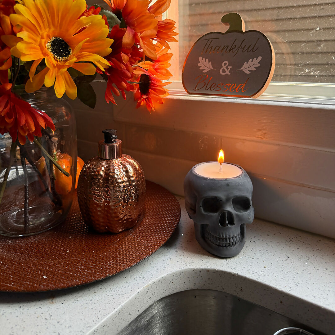Skull-shaped candle with a lit flame on a windowsill with flowers and a decorative sign.