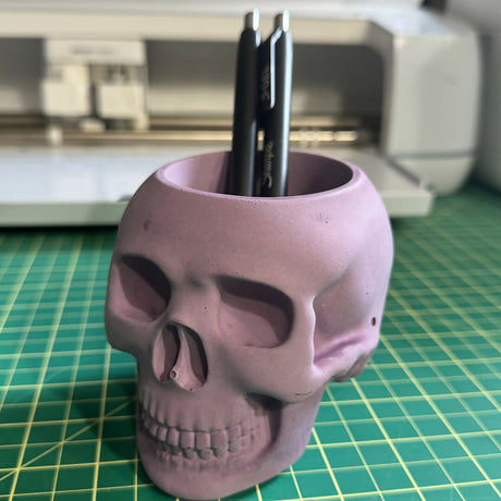 skull shaped candle holder holding pens on a green cutting mat in front of a cutting machine