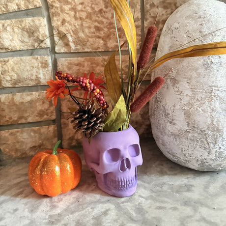 skull shaped candle holder with artificial foliage beside a small decorative pumpkin and large vase on a textured surface