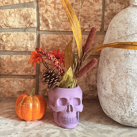 skull shaped candle holder with artificial foliage beside a small decorative pumpkin and large vase on a textured surface